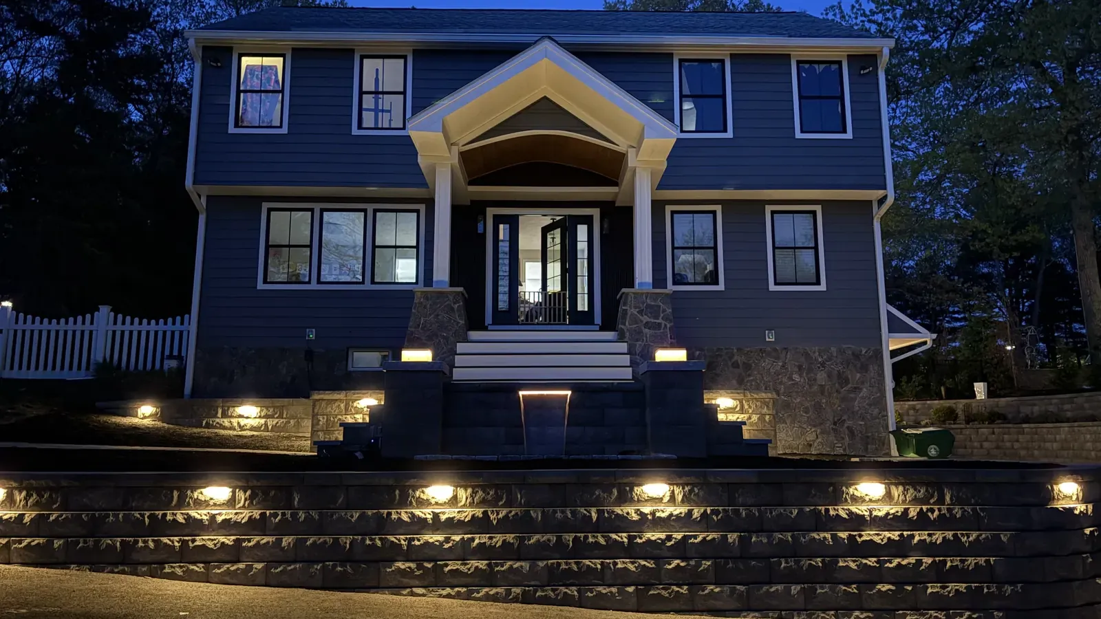 Block retaining wall with built-in lights and stone steps in front of a home at dusk